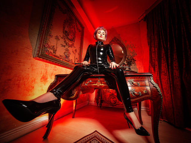 IsabelaConte in black latex outfit, sitting on wooden table, legs spread, high heels, red lighting, short pink hair, serious expression, hands on table, feet up, sexy.