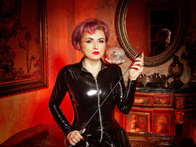IsabelaConte with short purple hair, wearing a shiny black latex zip-up, holding a riding crop, red lipstick, and a confident expression, in a dimly lit, red-hued room.