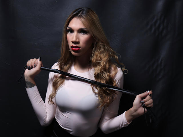 Littlebrunettehot with long blonde hair, red lipstick, white tight top, holding black leather whip, serious expression, dark background, right hand gripping whip, left hand near hip.