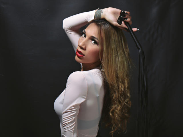 Petite blonde avec de longs cheveux blonds ondulés, rouge à lèvres rouge, haut blanc à manches longues, tenant un fouet en cuir noir, regardant par-dessus son épaule, fond noir, bras droit levé, tatouage au poignet..