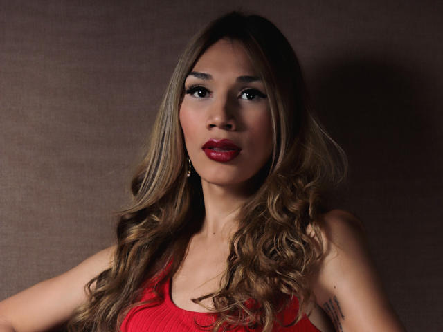 Une jeune femme brune, avec de longs cheveux bruns ondulés, du rouge à lèvres et un débardeur rouge, mettant en valeur sa poitrine et un tatouage sur son bras droit. Fond brun..