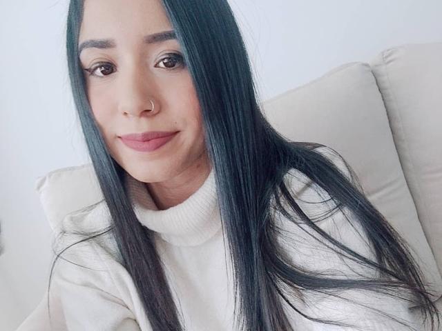 AshleyLover hat langes, schwarzes Haar, braune Augen, einen Nasenring und trägt ein weißes Rollkragenpullover. Sie lächelt sanft mit rosafarbenen Lippen. Ihre Brüste sind nicht sichtbar..
