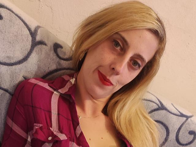 CoquineHots tiene el cabello rubio, lápiz labial rojo y lleva una camisa rosa de cuadros. Está acostada con una sonrisa sutil, mirando a la cámara. Sus pechos son pequeños..