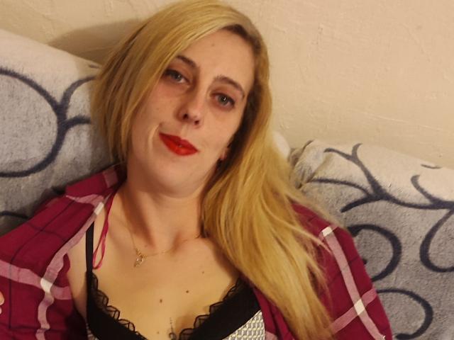 Una mujer atractiva con cabello rubio, lápiz labial rojo, vestida con una camisa rosa a cuadros, un sujetador de encaje negro, acostada sobre un cojín estampado, sonriendo a la cámara..
