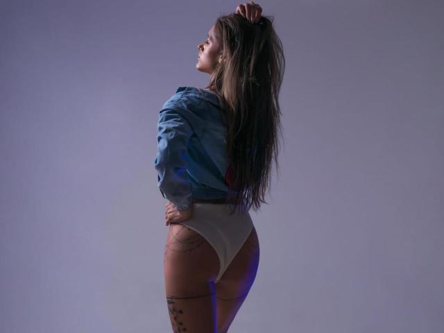 ElianMoon, con lunghi capelli castani, indossa una camicia blu e biancheria intima bianca, in piedi di profilo, con una mano tra i capelli, mostrando il sedere e la coscia tatuata..