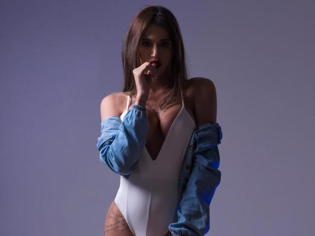 ElianMoon with brown hair, wearing a white one-piece, blue jacket off-shoulder, hand near mouth, tattoos on right hip, looking seductively at camera.