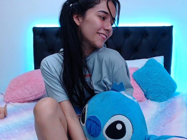DeisyWest with long black pigtails, smiling, wearing a gray tee, sitting on a bed with a blue Stitch plushie, pink and blue pillows, and a black tufted headboard.