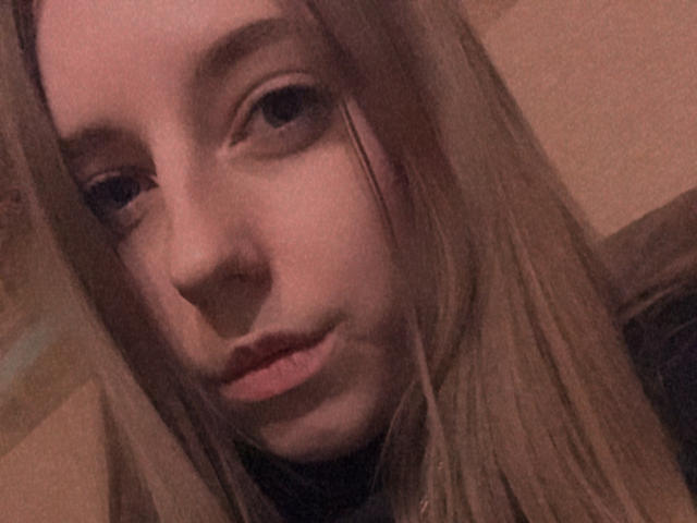 Het gezicht van BelleMartha, met lichtbruine ogen, steil blond haar en een lichte, rozeachtige glimlach, met een zachte, bruinachtige, vage achtergrond. Haar neus en lippen zijn gecentreerd..