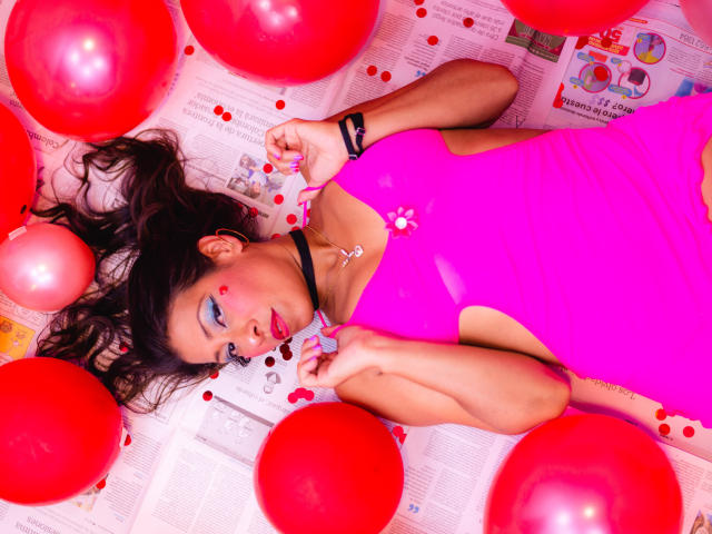 Lauren Hills acostada sobre globos rojos, vistiendo un vestido rosa brillante, con un punto rojo en la mejilla, sosteniendo un objeto con forma de corazón, mirando hacia arriba con ojos azules..