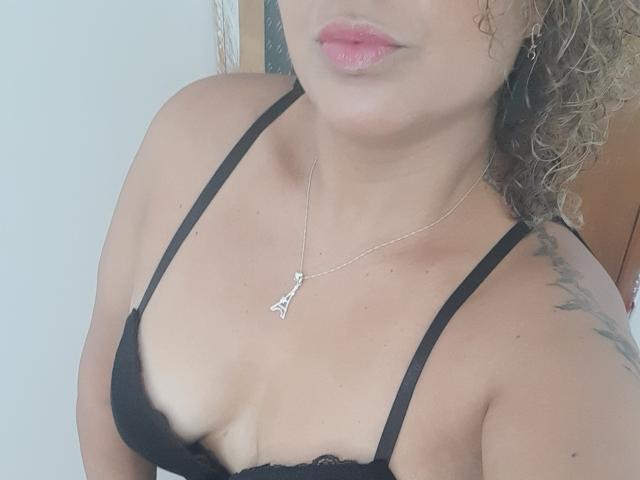 SolmaryHot with curly brown hair, wearing a black lace bra, silver necklace with a triangle pendant, and pink lipstick, showing her tits and a tattoo on her right shoulder.