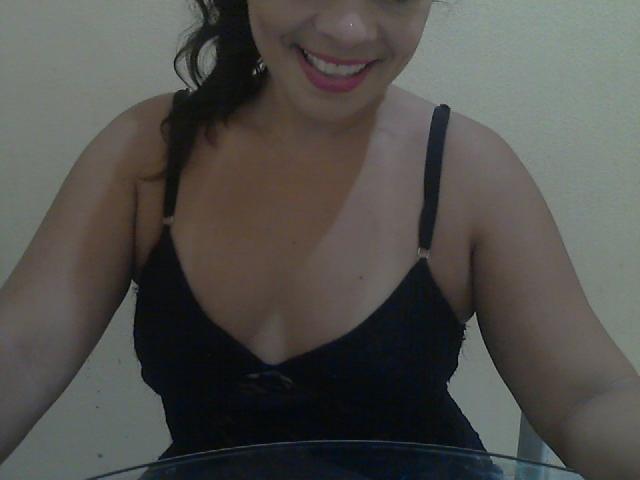 LauraPortela with dark hair, smiling, wearing a black spaghetti-strap dress that shows her tits. She's sitting at a table, upper body visible, hands out of frame.