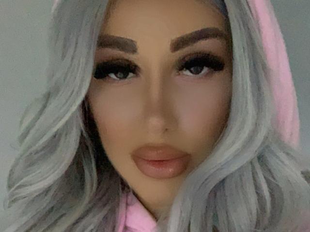TanyaMonroe with gray wavy hair, thick eyebrows, long black lashes, and pink hoodie, looking directly at the camera with a subtle smile. Her tits aren't visible.