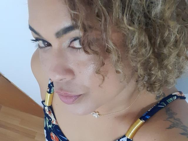 SolmaryHot with curly blonde hair, shiny skin, gold necklace, floral top, and gold straps, looking up with thick eyebrows and long eyelashes, showing a tattoo on her shoulder.
