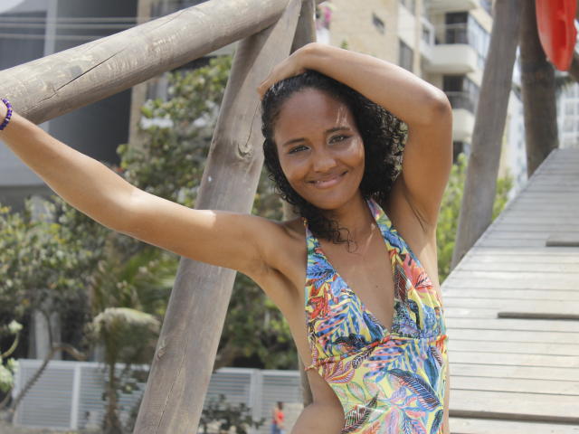 Megan Gil, con i capelli neri ricci, sorridente, indossa un top bikini colorato e a fantasia, con spalline, braccia alzate, mani su una struttura di legno, in piedi su una scala di legno, in una giornata di sole, con occhi verdi, pelle marrone media..
