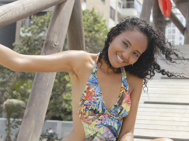 Megan Gil, con i capelli neri ricci, sorridente, indossa un bikini colorato, a taglio basso e con motivi, che mette in risalto il suo seno piccolo, sodo e prosperoso, seduta su una struttura di legno per bambini, sotto cieli sereni e soleggiati..