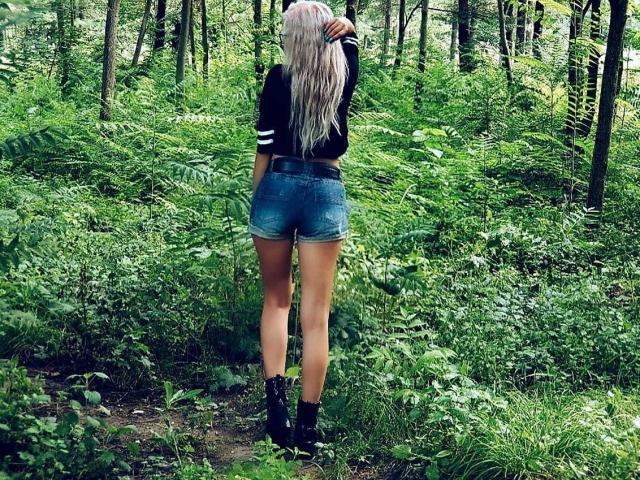 NaugtyBlonde stands with her back to the camera in a forest, wearing a black top, denim shorts, and black boots, with long, wavy blonde hair, and a visible ass.
