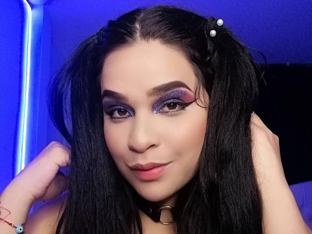 AntonelaPark with dark hair in pigtails, purple eyeshadow, thick eyebrows, pink lipstick, and a subtle smile, wearing a black choker, touching her hair, blue-lit background.