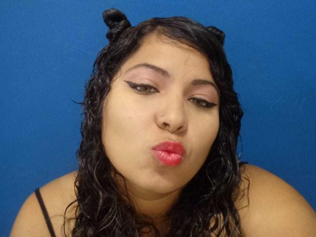 ArtemisaRoses tem cabelo preto e liso, olhos castanhos, sombra rosa, delineador preto, batom vermelho, um piercing no nariz e está fazendo um biquinho com os lábios, com um fundo azul..