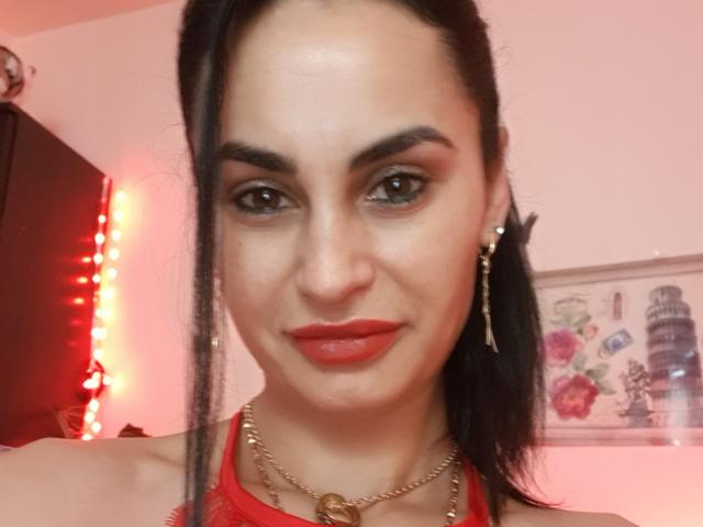 TheSunshine a les cheveux noirs attachés en queue de cheval, du rouge à lèvres, des boucles d'oreilles dorées, et un haut rouge à encolure américaine avec un collier en or, révélant un visage séduisant avec des yeux rouges..