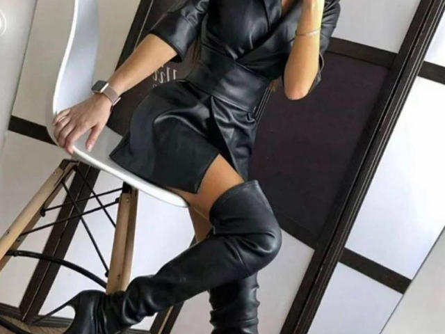 AliaAnita wearing a black leather dress with a side slit, black knee-high boots, and a wrist bracelet, sitting on a white chair with one leg crossed.