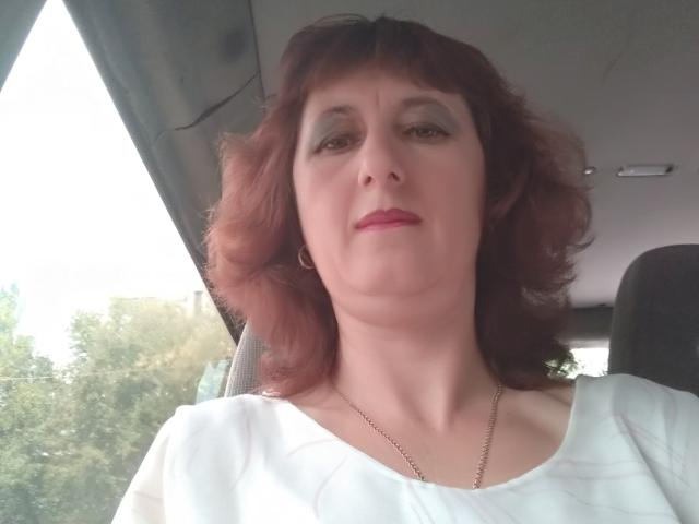 BellisimaAh ha i capelli rossi, la pelle chiara e indossa una maglietta bianca. È seduta in macchina con un sorriso appena accennato, guardando leggermente verso l'alto la fotocamera..