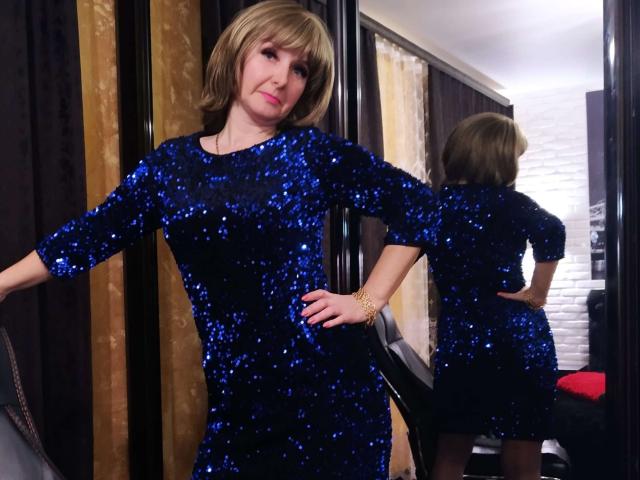 VAlica, a blonde woman with a cute face, wears a blue sequined dress, standing with arms out, reflected in a mirror, showing her ass and back.