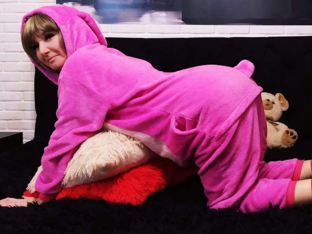 VAlica in a pink, furry, bear-themed onesie, kneeling on a black bed with a red and beige pillow, smiling at the camera, with a stuffed bear on her right.