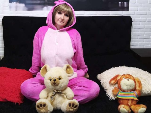 VAlica in a pink and white teddy bear pajama suit, sitting on a black couch, with a white teddy bear and orange bunny stuffed toy on either side.
