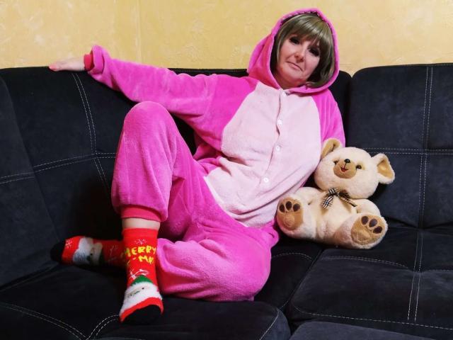 VAlica in a pink fluffy pajama onesie with hood, sitting on a black couch, wearing red socks with Christmas patterns, holding a beige teddy bear with a checkered bow.