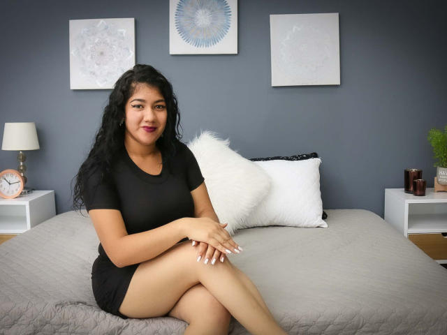 Kendall Owens mit lockigem, schwarzem Haar, trägt ein eng anliegendes, schwarzes Kleid und sitzt auf einem grauen Bett, die Beine gekreuzt, mit einem weißen Kissen im Rücken, an einer blauen Wand mit Kunstwerken, lächelnd, mit pinkem Lippenstift..