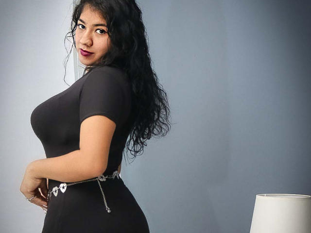 Kendall Owens, com cabelos pretos cacheados, vestindo uma blusa preta justa e um cinto com correntes, exibindo seu grande bumbum e cintura fina, olhando por cima do ombro com batom vermelho escuro..