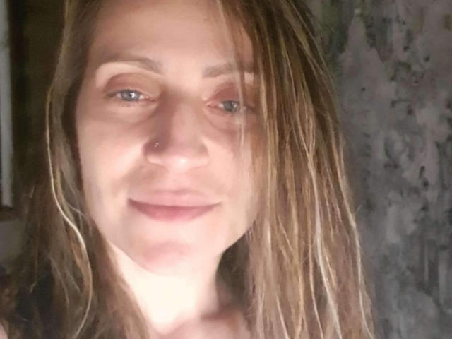 AliaAnita has light brown, wet, shoulder-length hair, blue eyes, a small nose piercing, and a subtle smile, with soft, natural lighting highlighting her fair skin.