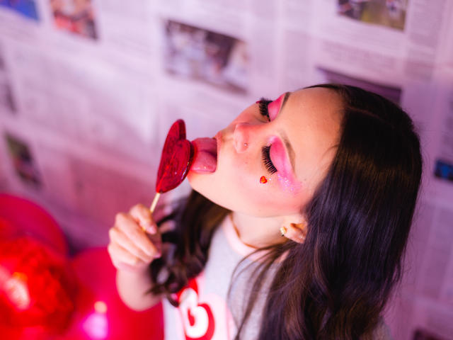 Jazmin Wells, com cabelo escuro, maquiagem rosa nos olhos e piercing no nariz, lambendo um pirulito vermelho em formato de coração, vestindo uma camisa branca com um desenho vermelho. Iluminação rosa e vermelha..