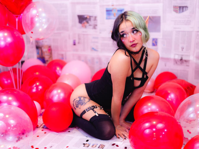 SalomePrince met groen en zwart haar, een zwart topje met bandjes, zwarte hoge kniebedekkers, een tatoeage op de rechter dij, knielend tussen rode en roze ballonnen, kijkend naar de zijkant, met een zwarte choker om..