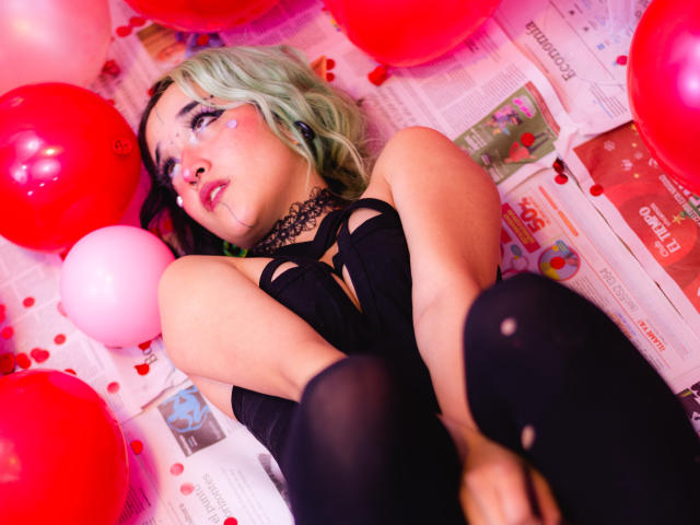 SalomePrince, com cabelo verde, gargantilha preta e blusa preta, cercada por balões vermelhos e rosa, deitada em um jornal com confetes em formato de coração, usando meias pretas, olhando para cima..