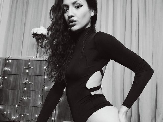 VievienneClemente with long curly hair, wearing a black cut-out leotard, showing her ass, standing, looking at the camera, with a vase and string lights in the background.
