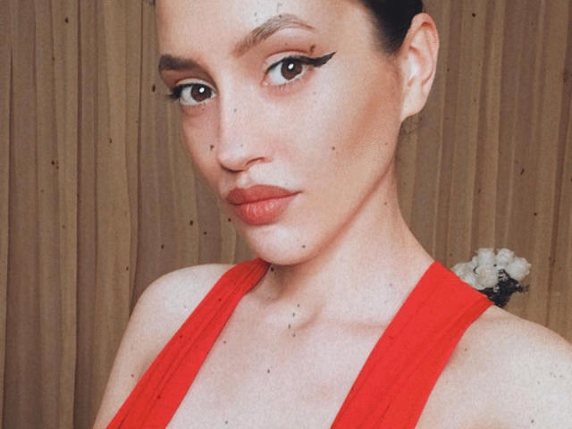 Vievienne Clemente mit dunklem Haar zu einem Dutt gebunden, trägt ein rotes, ärmelloses Kleid, auffälligen Eyeliner, helle Haut mit Sommersprossen, rote Lippen und eine kleine Blume im Haar..