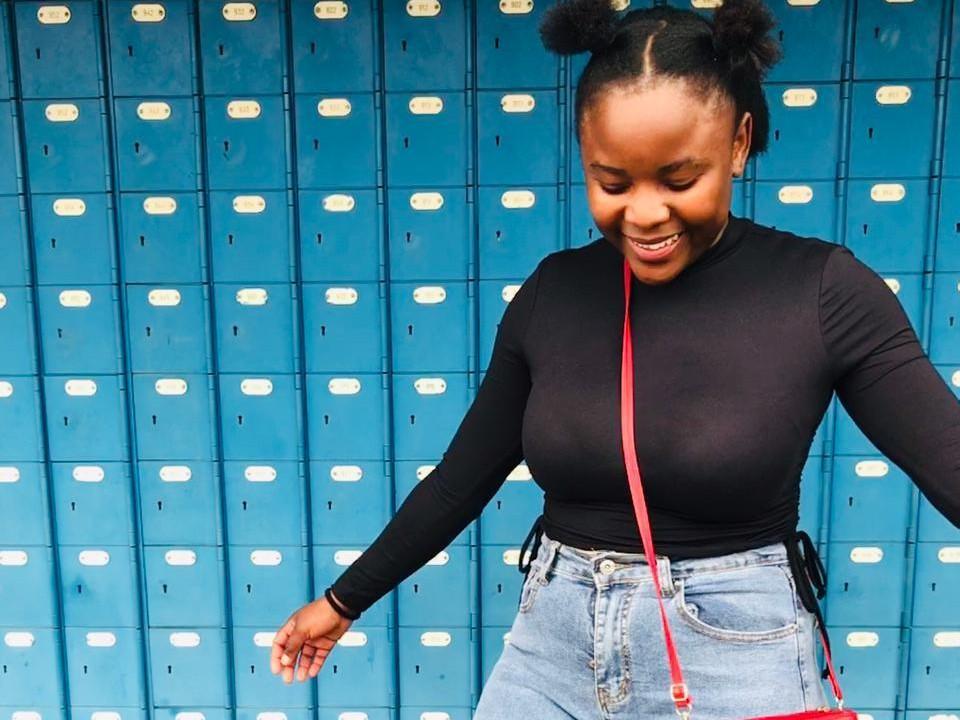 Una ragazza di nome HotXTamya, con la pelle scura, acconciatura con due chignon neri, top nero a maniche lunghe che mostra il seno, jeans blu a vita alta, bretella rossa, sorridente, in piedi davanti a degli armadietti blu..