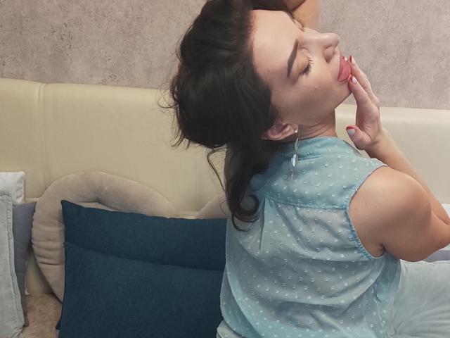 SimoniGrace, avec des cheveux foncés, les yeux fermés, la langue tirée, léchant son doigt, portant un haut bleu à pois, assise sur un lit beige avec des coussins bleus et beiges..