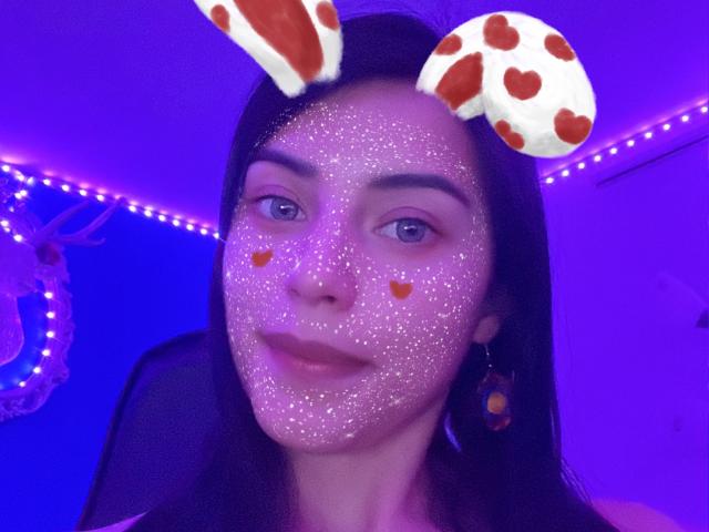 Une jeune fille avec des paillettes sur le visage, un emoji d'oreilles de vache, des emojis de cœurs orange sur les joues, un éclairage LED violet, des cheveux noirs, des yeux bleus et une peau teintée de violet dans un autoportrait..