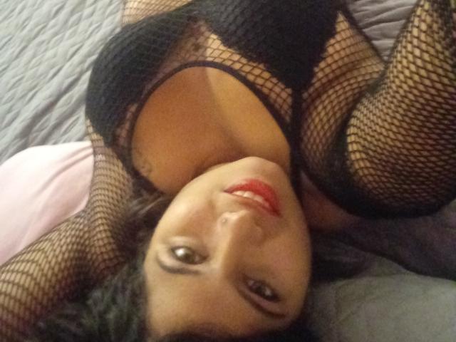 ArtemisaRoses está deitada em uma cama, vestindo lingerie de rede preta, com os seios à mostra, batom vermelho e uma tatuagem no lado direito. Seu rosto está de cabeça para baixo..