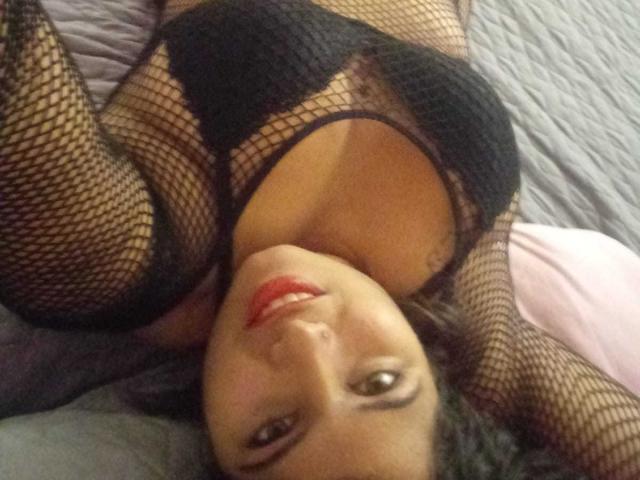 ArtemisaRoses, acostada en la cama, vestida con lencería de malla negra con los pezones a la vista, sonriendo con lápiz labial rojo, ojos marrones y cabello oscuro, mirando hacia arriba a la cámara..