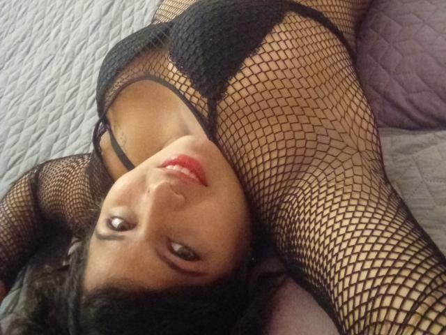 ArtemisaRoses liegt im Bett, trägt schwarze Netzstrümpfe, zeigt ihre Brüste, mit erhobenen Armen, blickt zur Kamera auf, trägt roten Lippenstift, hat dunkles Haar und lächelt..