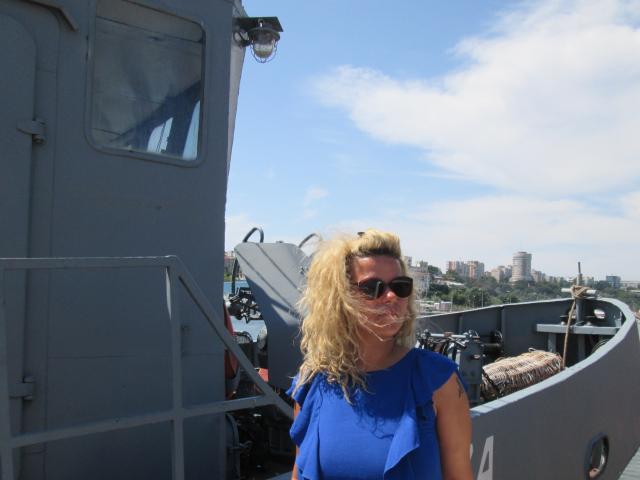 FacialEjac hat zerzaustes blondes Haar, trägt ein blaues Rüschenhemd und eine schwarze Sonnenbrille und steht auf einem grauen Schiff, im Hintergrund sind ein Motor und Seile zu sehen..