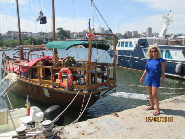 A wooden sailboat with a green canopy and life ring, moored beside a dock. FacialEjac stands in a blue dress, holding a phone, with a boat in the background.