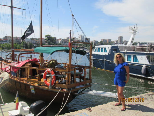 Blond FacialEjac in blue dress stands next to wooden pirate ship with flag, life ring, and rope, boat in background, wearing red sandals, on sunny day, watermark visible.