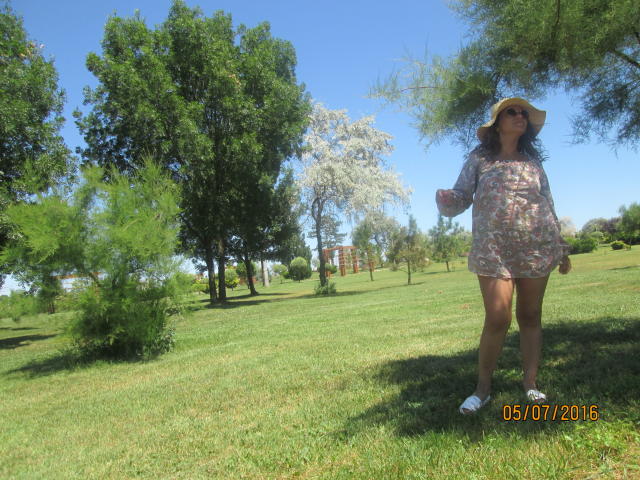 FacialEjac has medium tits, wears a floral dress, sunhat, and white sneakers, standing in a grassy park with trees, holding her arm out, and a blue sky.