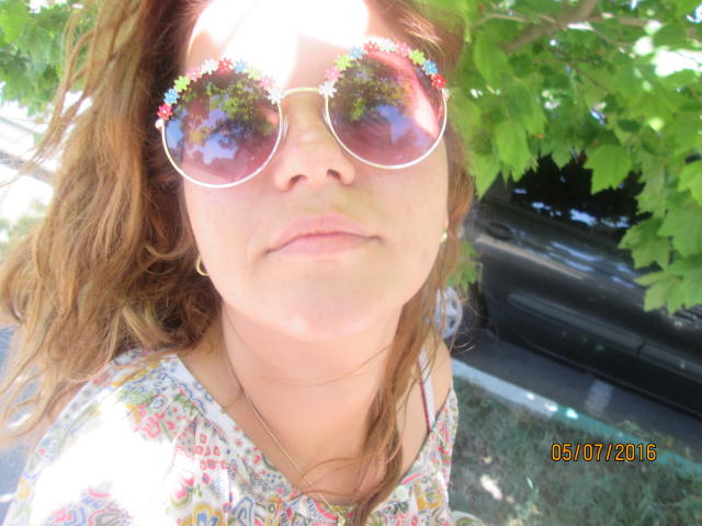 FacialEjac with reflective sunglasses, floral dress, and wavy brown hair, showing a subtle, closed-lip smile. Her tits are small and her nose is slightly freckled.