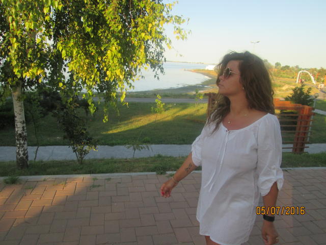 FacialEjac hat welliges, braunes Haar, trägt eine Sonnenbrille und ein lockeres, weißes Kleid und steht auf einem Ziegelweg, blickt zur Seite, wobei sich im Hintergrund ein Baum und eine Rasenfläche befinden..