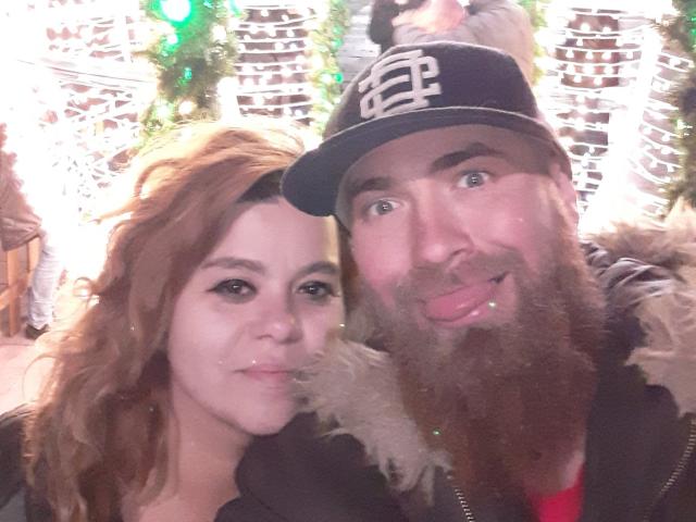 Man met baard en zwarte pet, die zijn tong uitsteekt, en een vrouw met golvend bruin haar, beiden glimlachend. Ze heeft kleine borsten, hij heeft een grote achterkant. Kerstverlichting op de achtergrond..